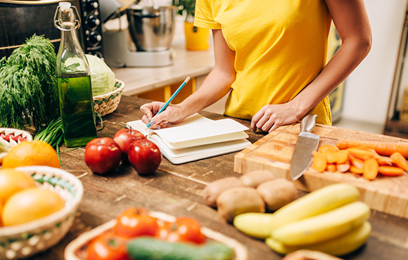 person writing in a notebook in a kitchen surrounded by fresh fruits and vegetables including apples tomatoes carrots and bananas focused on meal planning and healthy recipes 9 ingredients and nutritional choices