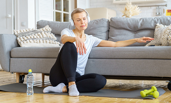 senior woman exercising at home wearing headphones sitting on a yoga mat with water bottle and dumbbells promoting fitness and health 2 ways to stay active