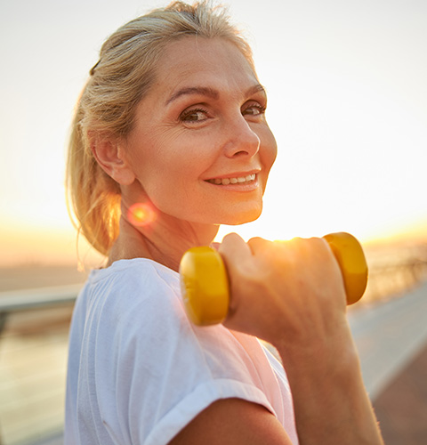 smiling woman holding a yellow dumbbell during sunset fitness and health routine 3 exercises